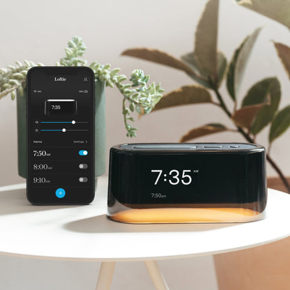 The  Tuft and Needle Loftie Smart Alarm Clock in a bedroom on a table next to a phone with the Loftie app